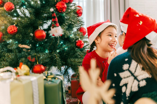 Happy mother and young daughter wearing Santa hats laughing together beside a decorated Christmas tree and wrapped gifts, sharing a loving and joyful moment during the festive holiday season.
