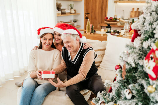 Family wearing Santa hats exchange a wrapped gift beside a decorated Christmas tree in a cozy living room, smiling and enjoying a warm holiday moment