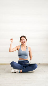 Smiling woman in activewear sits cross-legged with a laptop, raising a celebratory fist during a home fitness session, suggesting online workout or virtual training against a minimal white wall.
