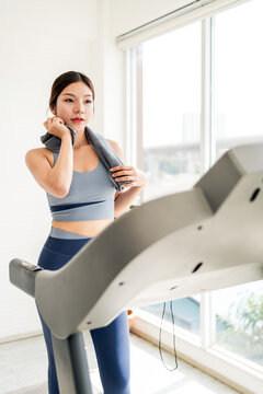 Woman in activewear cooling down on a treadmill, holding a towel in a bright home gym with large windows and natural light.