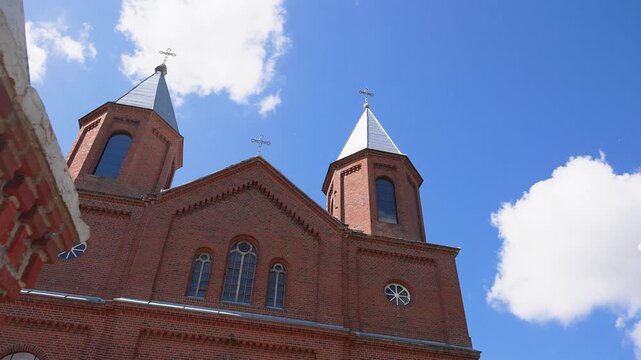 Striking low angle footage pans upward, revealing red brick church towers under a vivid blue sky with scattered clouds. Sunlight highlights the architectural detail in this steady, wide angle shot.