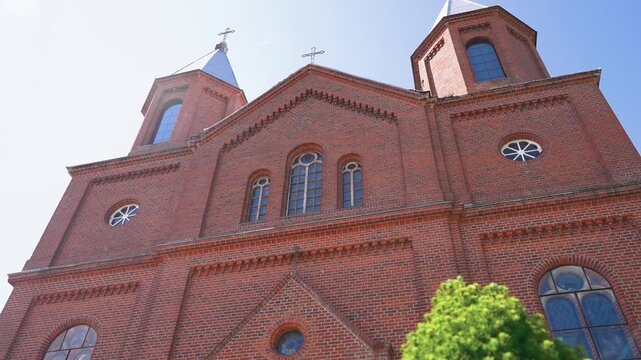 Low-angle shot of a striking red brick church facade with twin towers and arched windows, captured in bright daylight. Sunlight highlights detailed architecture, vibrant atmosphere, and clear blue sky