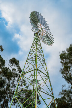 Low angle view of a traditional windmill with its weathered green steel lattice tower and metal blades rising against a bright, cloud-filled blue sky, Donald, Victoria, Australia