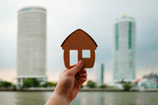 Real estate and waterfront property concept with hand holding wooden house model against river and modern high rise condominium buildings for urban luxury living, apartment and home buying.