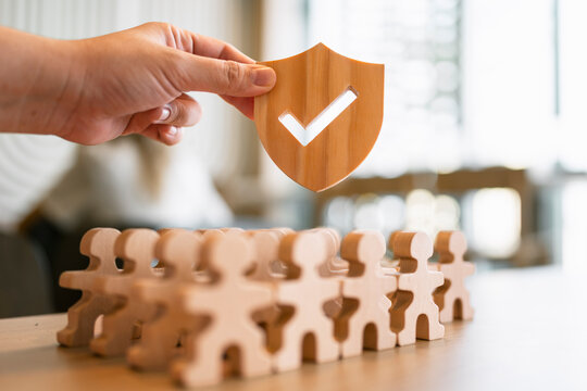 Life insurance and employee protection concept with hand holding wooden shield with checkmark over people figures for healthcare coverage, social security guarantee and risk management.
