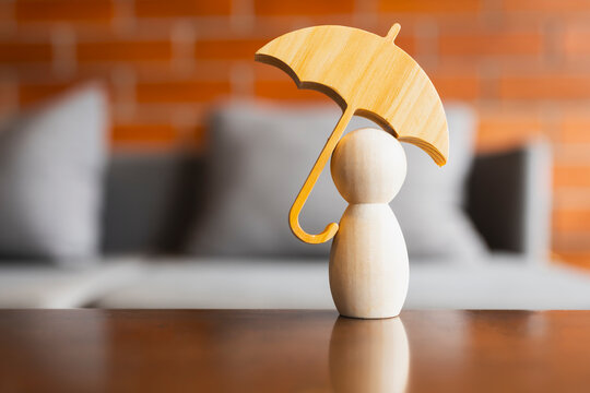 Life insurance and personal safety concept with single wooden peg doll figure standing under umbrella on reflective table with brick background for healthcare protection and security.