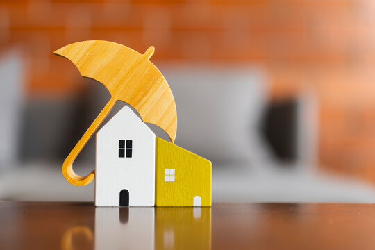 Home insurance and property protection concept with wooden umbrella covering miniature houses on reflective table with brick background for real estate mortgage, safety and risk coverage.