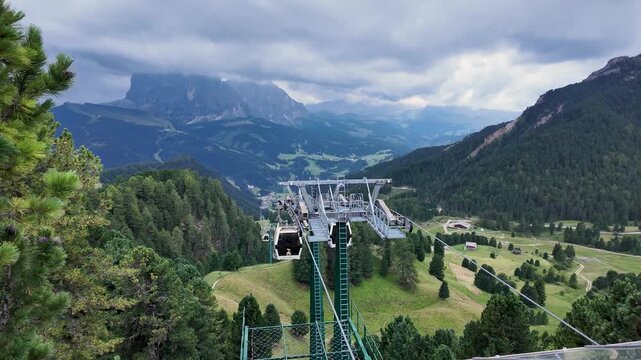 Scenic view of the Col Raiser cable car station above Seceda, surrounded by alpine meadows and forested slopes, with dramatic Dolomite mountains and low clouds stretching across Val Gardena