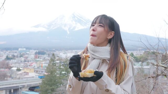 Young Asian woman tourist eating local street food with Mount Fuji view in Japan. Female traveler enjoying Japanese snack during winter sightseeing, holiday vacation and cultural travel experience.