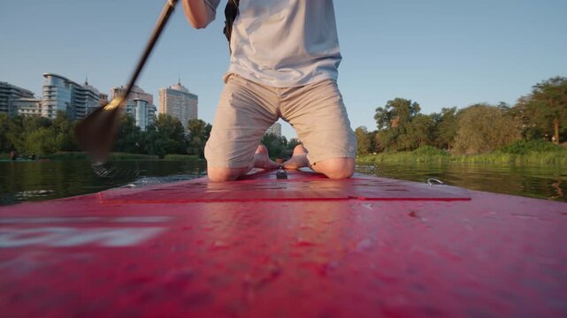 Man Kneeling on Red Paddleboard Paddling on River in City | Person Rowing SUP Board Toward Urban Skyline on Summer Morning | Active Outdoor Water Recreation Lifestyle