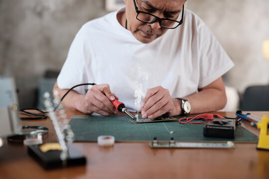 Mature Asian man repairs home electrical appliance, disassembling and soldering circuit board, focusing on precision detail, DIY fix tools, electronics component workshop hobby, expert handyman.