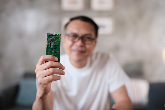 Selective focus on circuit board showing a mature Asian man proud of a successful soldering job, cheerful job achievement, home electronics appliance repair handyman, DIY fix tools, hobby workshop.
