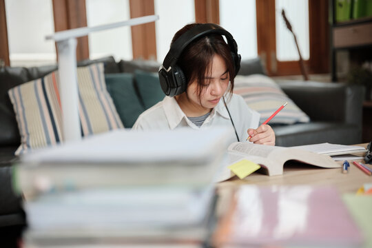 Asian teenage girl in headphones concentrates on learning at home, reading textbooks, note writing, self-tutorial exams, preparation for back to school, Homeschooling, university education lifestyle.