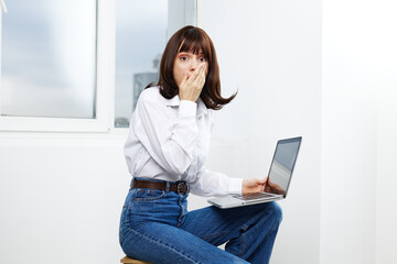 Young woman sits on a chair with a laptop, reacting with surprise as she covers her mouth. A...