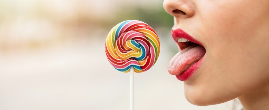 Woman licks a round rainbow lollipop showing her red lips and tongue clearly.