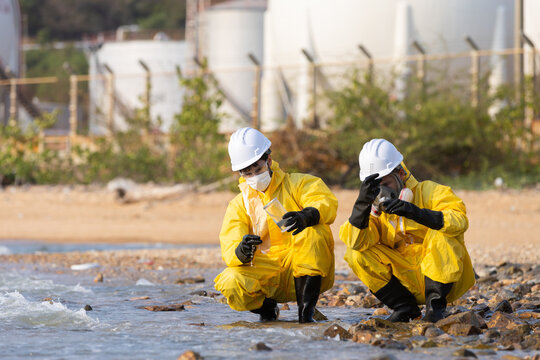 Serious environmental worker in hazmat suit inspecting polluted water at river edge alongside safety inspector with focused expression during daytime hazardous material spill testing