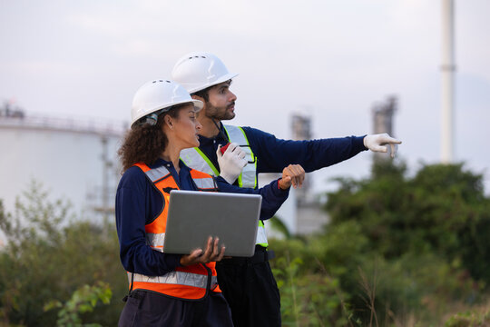 Industrial engineers wearing hard hats and safety vests using laptop and radio at oil facility, representing teamwork, inspection, communication, engineering and workplace safety.