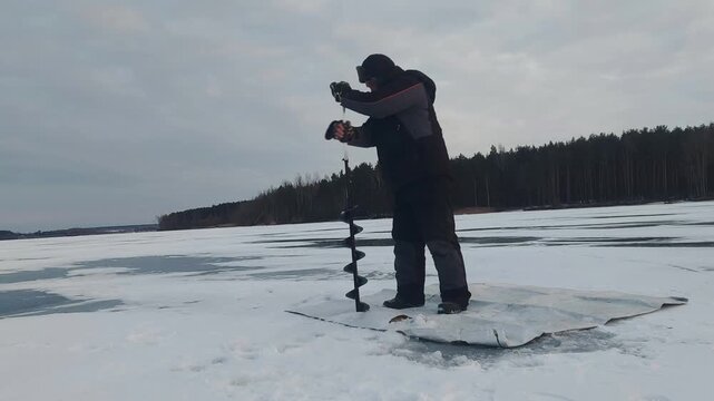 A person stands on a snowy surface, using an ice auger to drill a hole in the ice of a lake during winter. The surrounding area features trees and a gray sky. This activity shows the process 