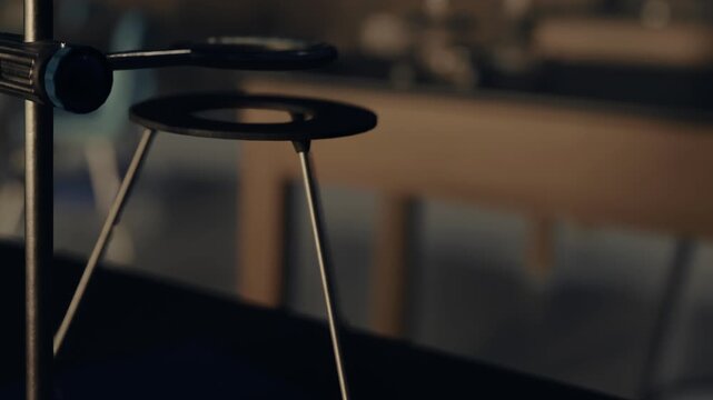 A stand is shown in focus, with a black ring positioned on top. The background shows an organized workspace with tools and equipment. Light from above highlights the stand.