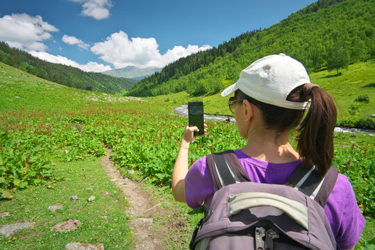 Female tourist with a backpack taking photos of mountains on her smartphone from a green valley path. Modern mobile travel blogging