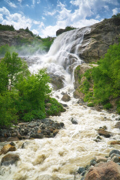 Powerful waterfall cascades down steep rocky cliffs surrounded by green trees under a cloudy sky. Alpine river stream fills travel brochures