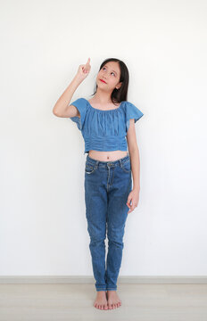An Asian preteen girl wearing a blue denim crop top and jeans stands against a white background, looking up and pointing her index finger upwards.