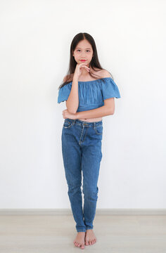 A young Asian girl wearing a blue off-shoulder crop top and jeans poses gracefully with her hand on her chin against a white wall.
