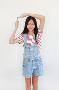 Asian girl in red and white striped t-shirt and blue denim overalls, standing and framing her face with both hands in a cute pose against white background.