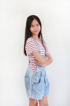 Asian preteen girl in striped t-shirt and denim dungaree shorts stands with crossed arms against white background, smiling gently at camera.