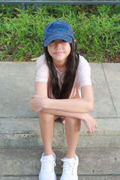 Portrait closeup of Asian girl, wearing denim cap and a light pink t-shirt, sits comfortably on a concrete floor in a garden setting, smiling gently towards camera.