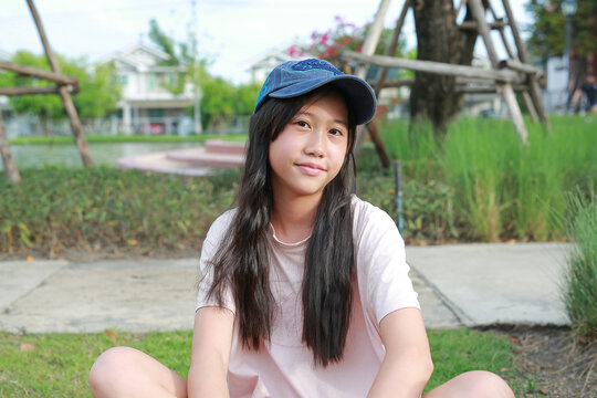 A young Asian girl wearing a blue cap and pink t-shirt is sitting on the grass in a park, smiling gently at the camera.