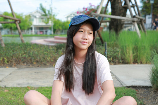 A young Asian girl wearing a blue cap and pink t-shirt sitting cross-legged on the grass in a park, smiling gently while looking to the side.