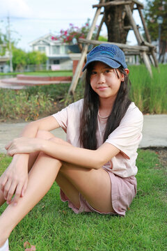 A young Asian girl wearing a denim cap and pink casual outfit sitting on the green grass outdoors in a park and smiling at the camera.