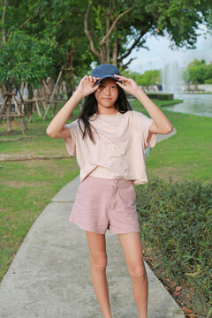 Portrait of young Asian girl standing on walkway in a park, holding her cap with both hands and looking at the camera.
