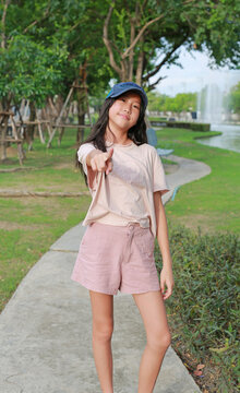 A young Asian girl wearing a cap and pink outfit is standing in a park, smiling and pointing her finger directly at the camera.