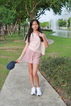 A young Asian girl posing with her cap in hand while standing on a concrete pathway in a green park.