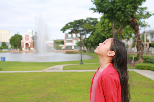 A young Asian girl standing in park with her eyes closed, tilting her head back to breathe in the fresh air and enjoy a peaceful, relaxing moment in nature.