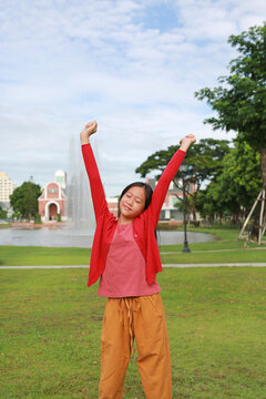 A young Asian girl standing in a park with raised arms and closed eyes, stretching outdoors on a bright sunny day.