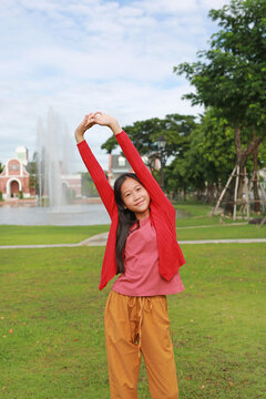 Happy young Asian girl is stretching her arms high up in the air while standing on the green lawn of a park, smiling cheerfully.