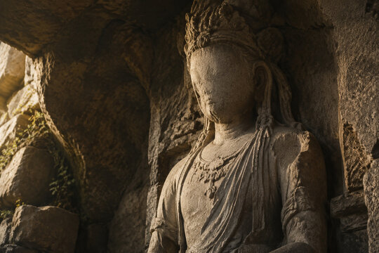 An ancient unfinished stone statue of a deity with a worn face in a rocky alcove.