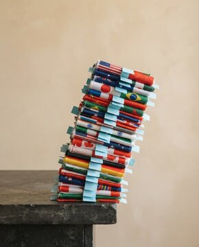 A precarious stack of rolled world flags with small blue paper tabs, leaning on a dark stone surface against a beige background with copy space for diplomacy campaigns