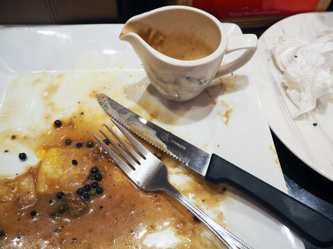 A messy plate after a delicious meal, with leftover gravy, peppercorns, a fork, and a knife. A small pitcher of gravy sits nearby, along with a crumpled napkin