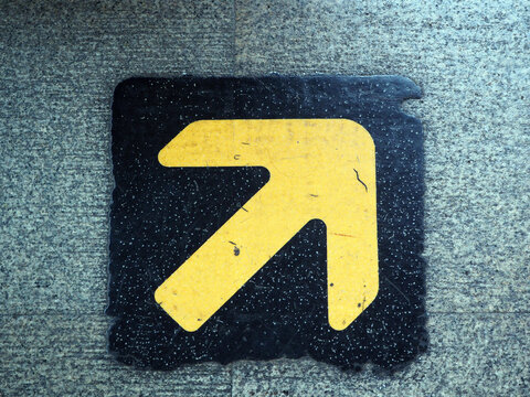 A yellow arrow on a black background points upwards and to the right, indicating direction. It is set against a textured grey surface
