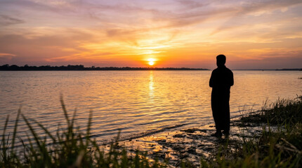 Fototapeta premium Silhouette man standing on muddy riverbank watching orange golden sunset sky over peaceful calm water during evening landscape.