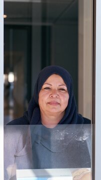 Latin American woman in a navy blue headscarf holding a prosthesis box, featuring a resolute expression behind a glass partition for health and survivorship covers