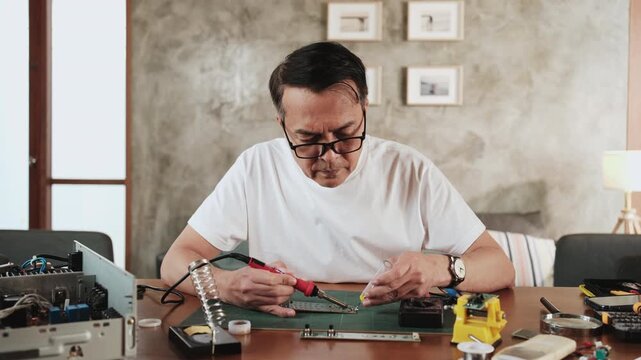 Mature Asian man repairs home electrical appliance, disassembling and soldering circuit board, focusing on precision detail, DIY fix tools, electronics component workshop hobby, expert handyman.