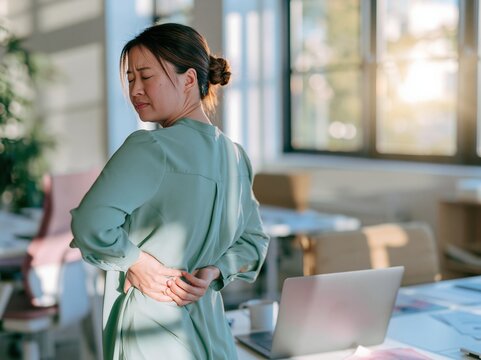 Asian woman in a grey blouse holding her lower back in a bright office with a laptop, showing physical strain for ergonomic education and workplace health