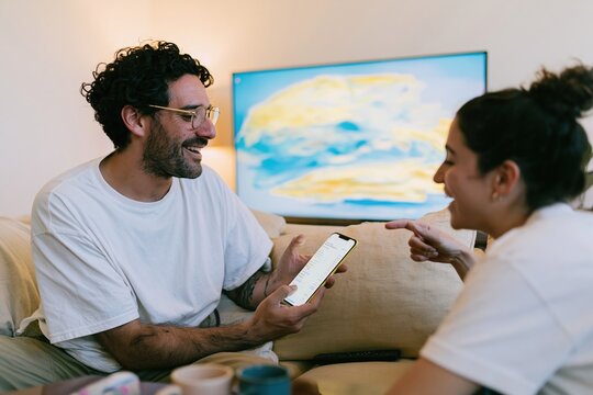 Hispanic man and woman sitting on a beige sofa, naturally discussing a phone screen with a billing list template for subscription management and finance