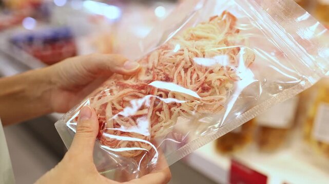 Close-up of a woman hands holding and choosing a vacuum-sealed plastic package of dried shredded squid or seafood snack from display shelves in a grocery store or supermarket. Retail shopping, food.