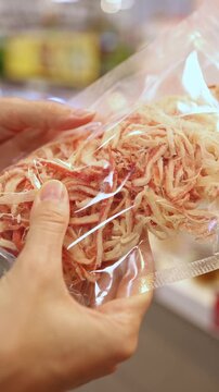 Close-up of a woman hands holding and choosing a vacuum-sealed plastic package of dried shredded squid or seafood snack from display shelves in a grocery store or supermarket. Retail shopping, food.